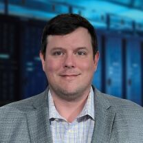 A man with dark hair and blue eyes smiles while wearing a grey blazer over a checkered shirt, with a blurred background