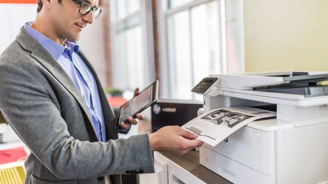 A man in a suit holds a tablet while retrieving a printed document from a printer.
