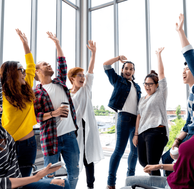 A diverse group of young adults celebrates with raised hands in a bright, modern office space.