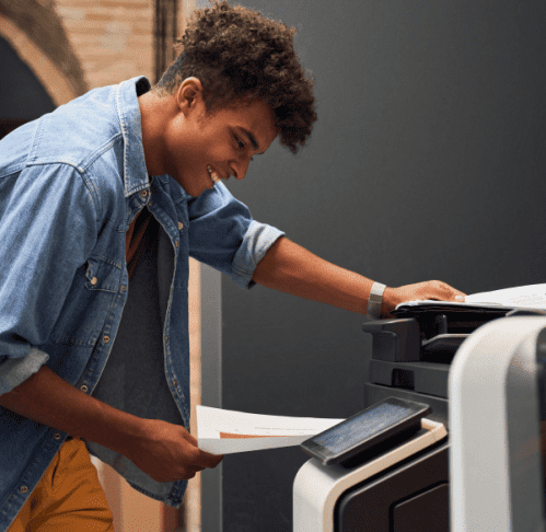 A young man in a denim jacket smiles as he places documents into a printer.