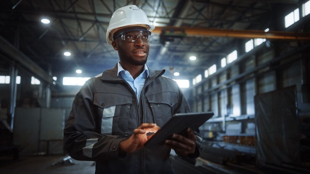 A Black man in a hard hat and safety glasses uses a tablet in a large industrial warehouse.