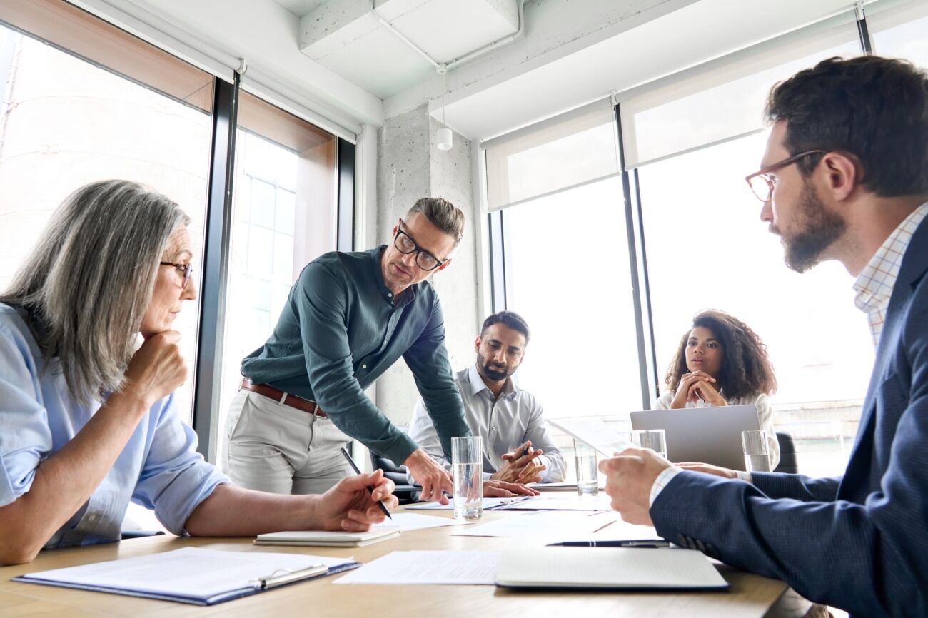 A diverse group of professionals are engaged in a meeting around a conference table, with one man pointing at documents.