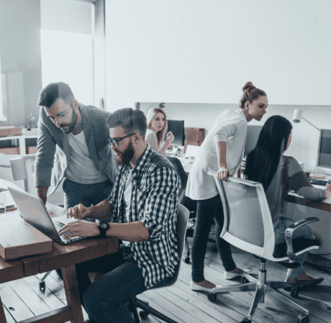 A group of colleagues collaborates in a modern office setting, with two men focused on a laptop and others working at