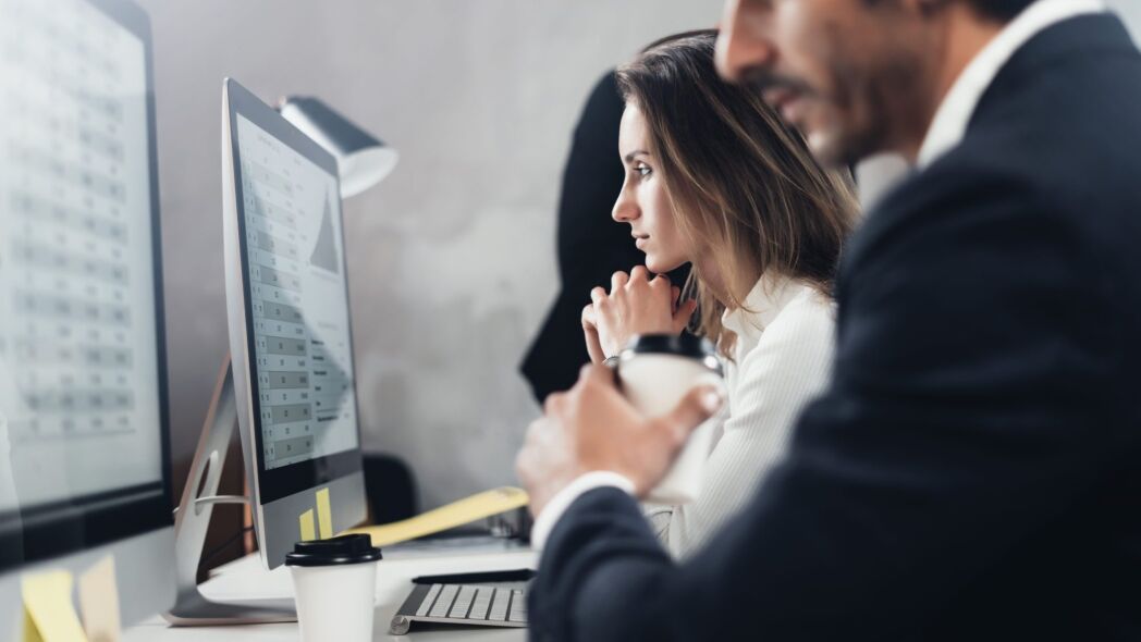 Two colleagues, a woman and a man, are working together at their computers in an office setting.