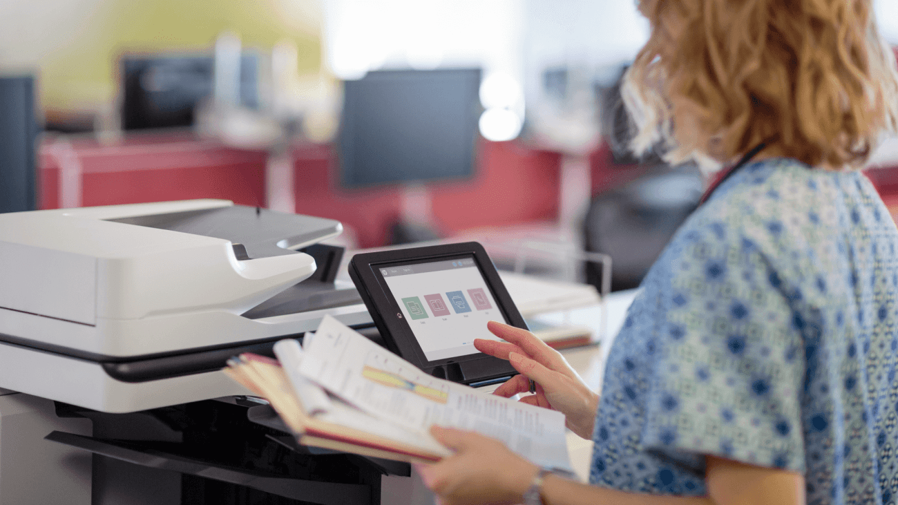 A person in a blue patterned shirt uses a touchscreen interface on a scanner to operate the device, holding a stack of
