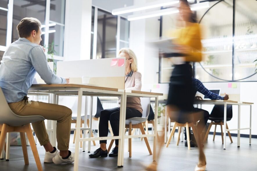 A blurred figure walks past a group of people working at desks in a modern office.