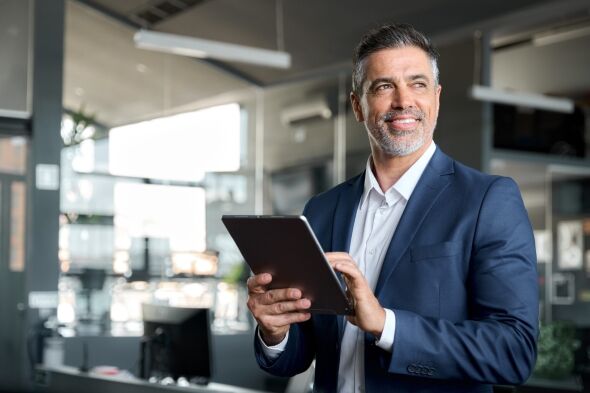 A smiling businessman in a navy suit holds a tablet and looks thoughtfully into the distance within a modern office