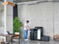 A man in a white t-shirt and jeans operates a large, modern office printer in a minimalist loft-style space with