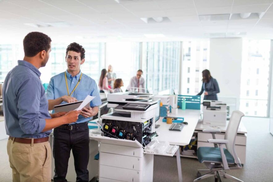Two men discuss documents near a disassembled printer in a modern office environment.