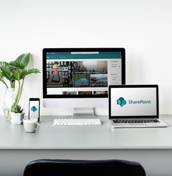 A modern workspace features a desktop monitor, laptop, and smartphone displaying the SharePoint interface, alongside a