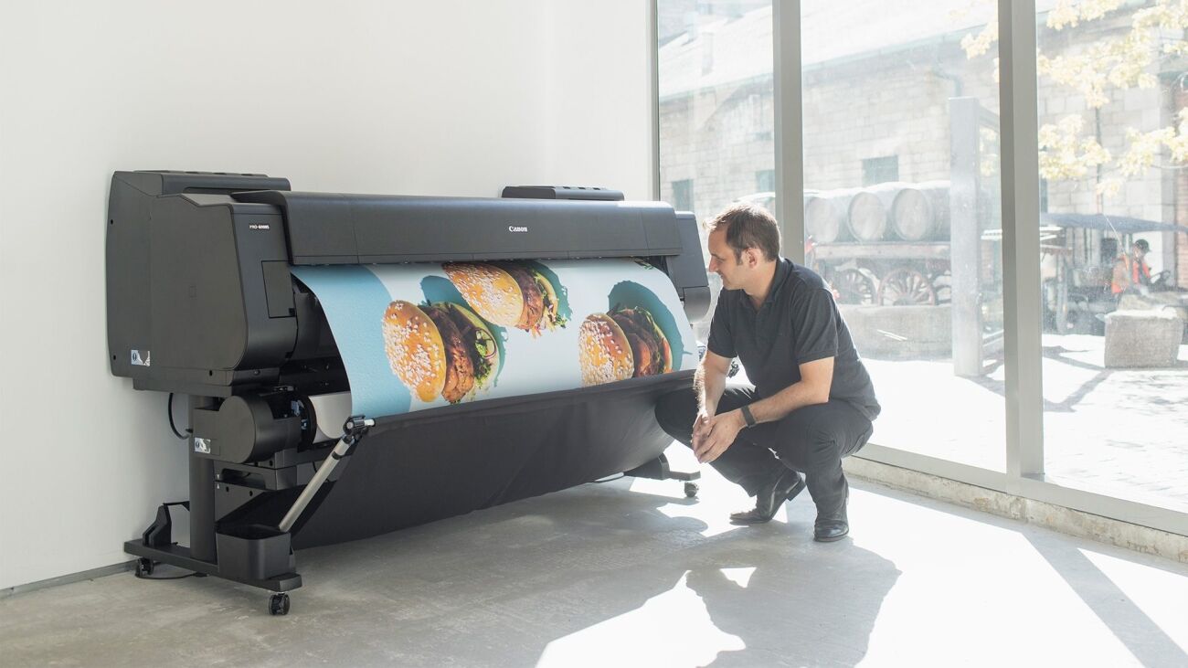 A man kneels to examine a large format printer displaying an image of burgers.