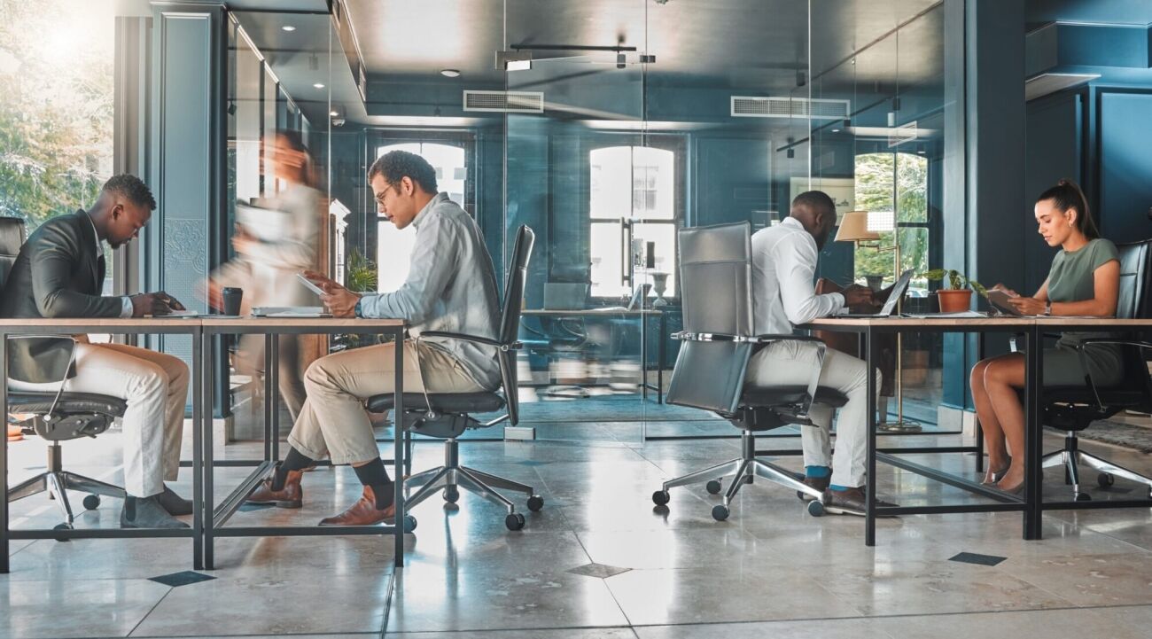 Four professionals work at desks in a modern office with glass partitions.