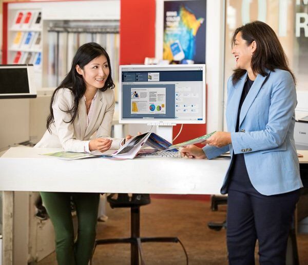 Two women in business attire are discussing documents and a computer screen in an office setting.
