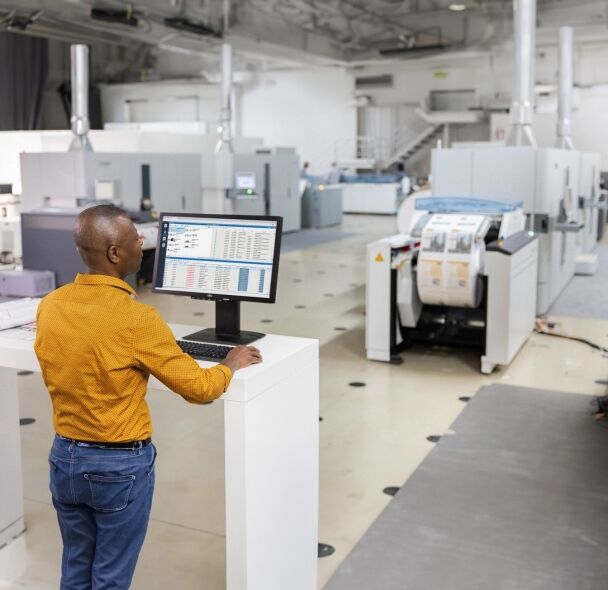A man in an orange shirt and jeans stands at a computer in a printing facility, overseeing operations.
