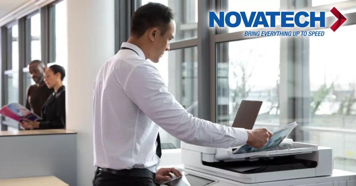 A man in a white shirt and tie operates an office printer, while two colleagues stand in the background and a Novatech