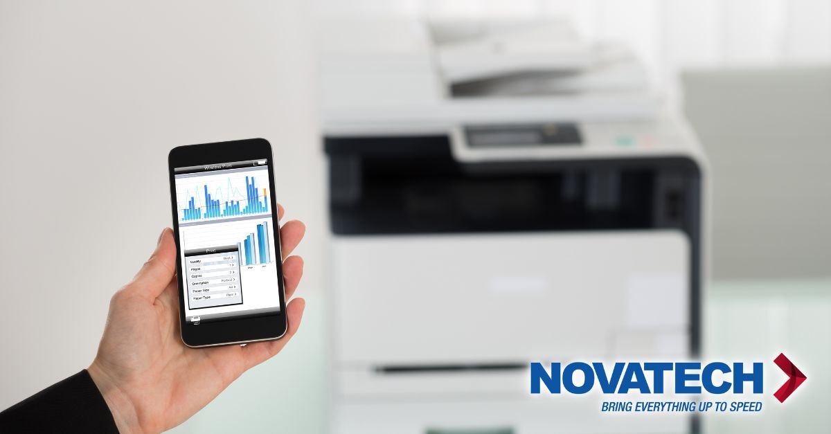 A hand holds a smartphone displaying charts and print settings, with a blurred office printer in the background and the