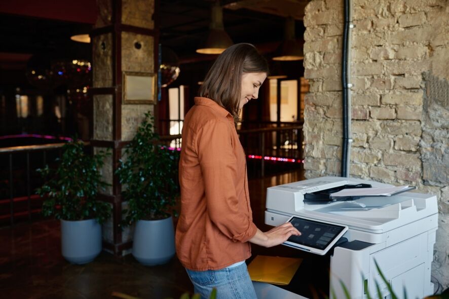 A smiling woman operates a modern multifunction printer within a warm, brick-walled interior.