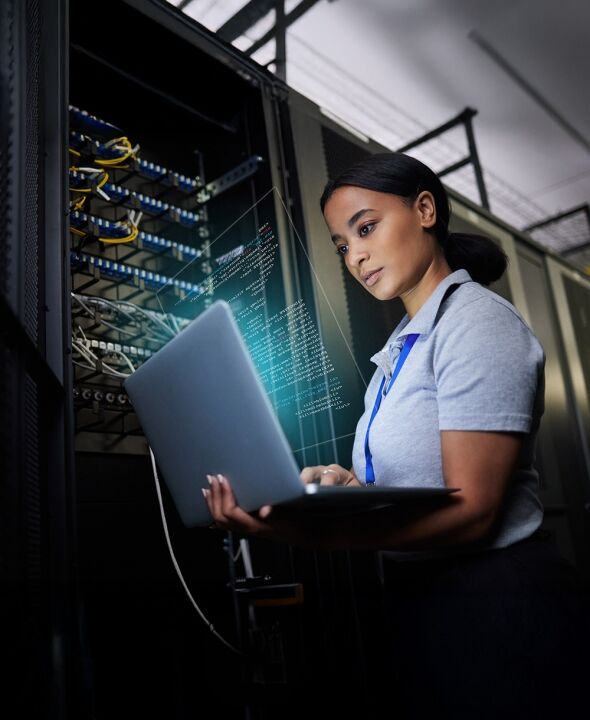 A woman in a data center works on a laptop, with code displayed on a transparent overlay.
