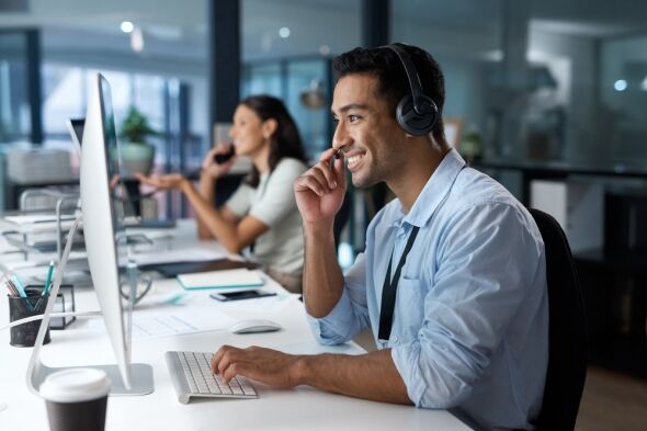 A man wearing headphones smiles while working at a computer in an office, with a woman on the phone in the background.