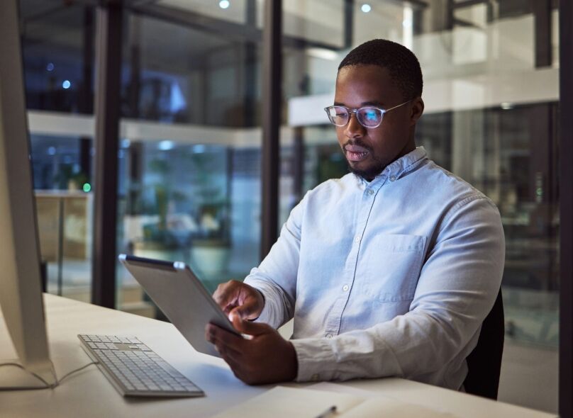 A man works diligently at a desk in a modern office at night, focused on a tablet in his hands.