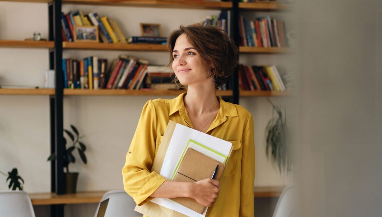 A young woman in a yellow shirt holds a stack of papers and a notebook while looking thoughtfully to the side in front