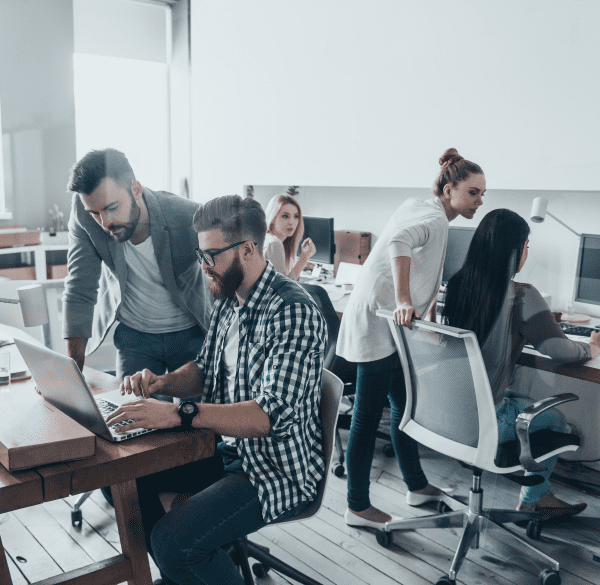 A group of colleagues collaborates in a modern office setting, with two men focused on a laptop and others working at