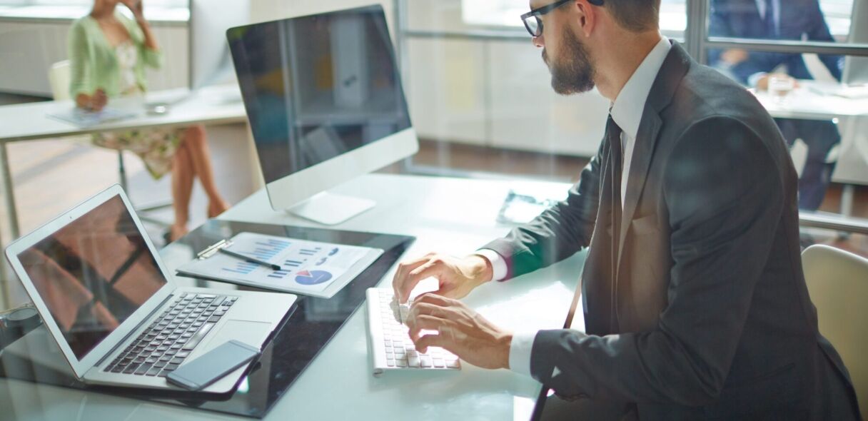 A man in a suit works on a laptop in an office setting, with a colleague in the background.