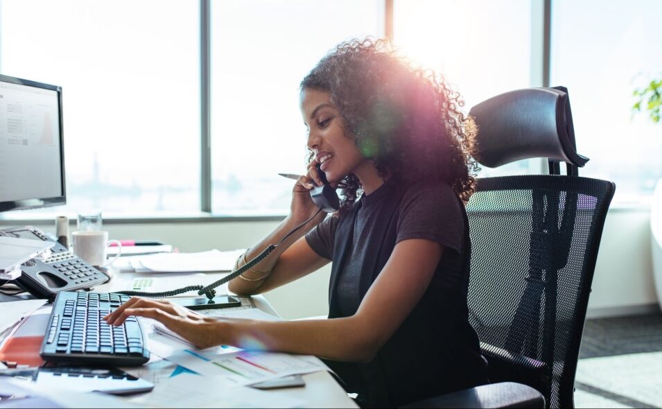 A woman with curly hair sits at a desk, typing on a keyboard and talking on a phone, with a bright window behind her.