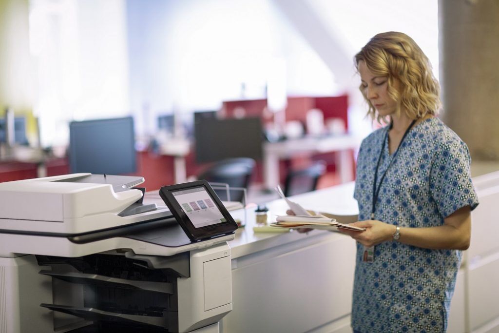 A woman in scrubs stands at a copier, holding a stack of papers.