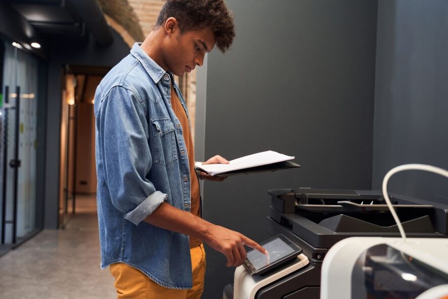 A young man in a denim shirt and orange pants uses a touchscreen on a copier while holding a stack of papers.