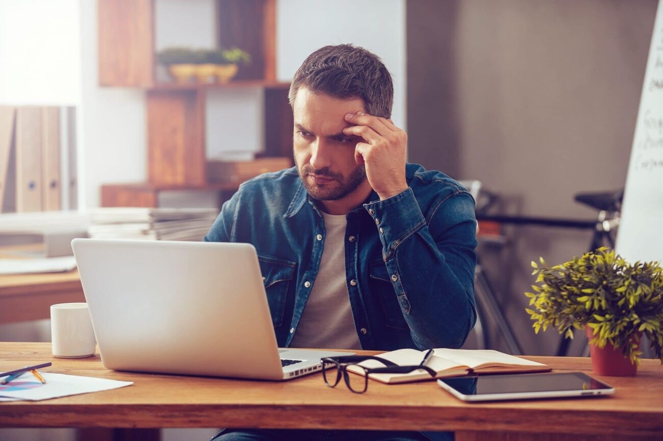 A man with a beard and short hair, wearing a denim shirt, sits at a wooden desk with a laptop, a notebook, glasses, and
