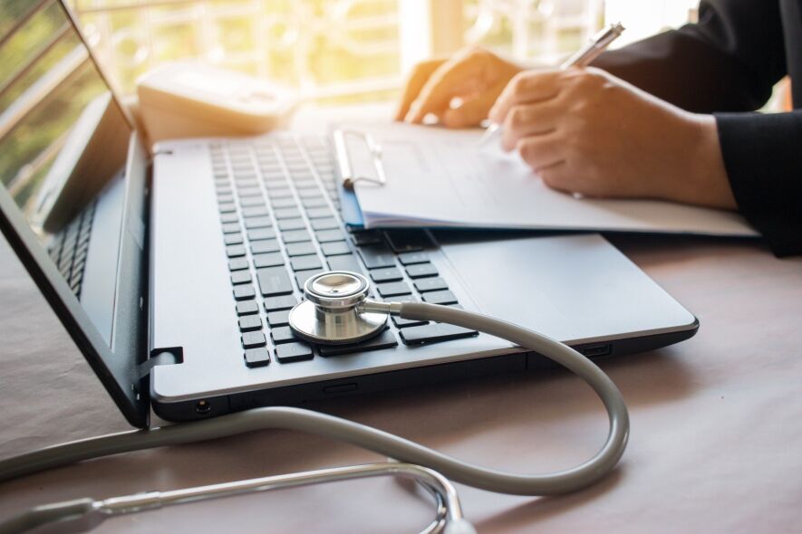 A stethoscope rests on a laptop keyboard as a person in the background writes on a document.