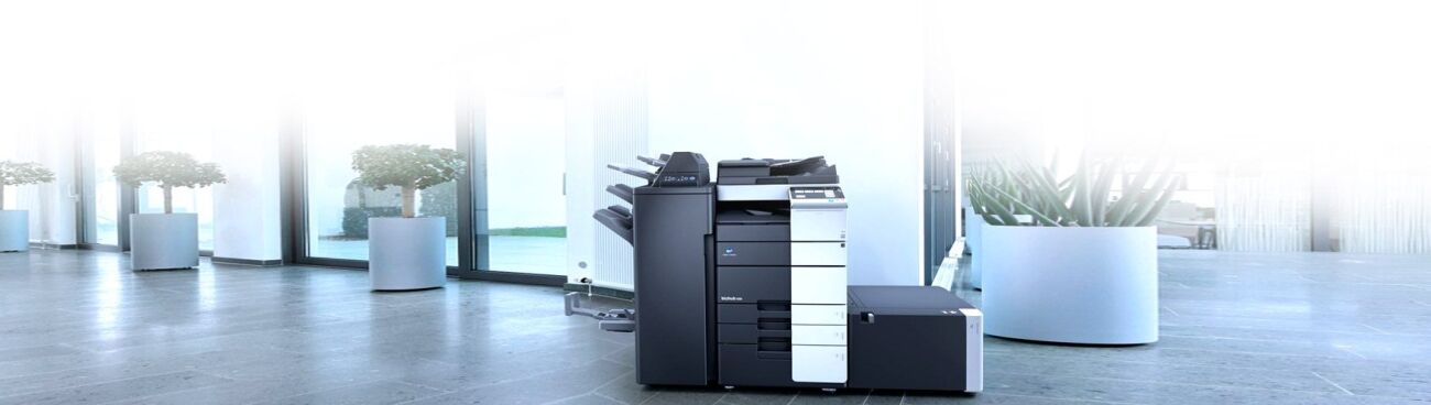 A modern office space features a large black and white multifunction printer, potted plants, and large windows.