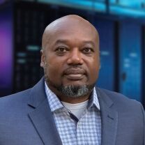 A man with a bald head and beard, dressed in a blue suit jacket and plaid shirt, stands in front of a blurred