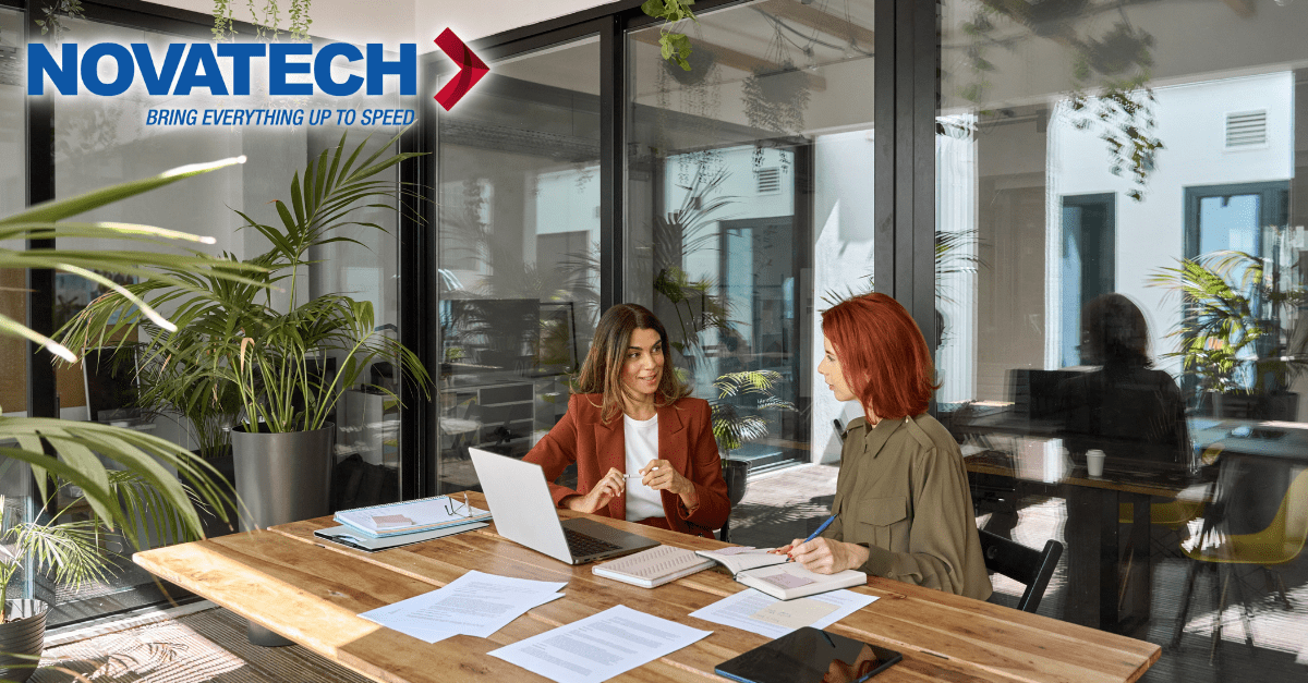 Two women are working at a table in a modern office with large windows and plants.
