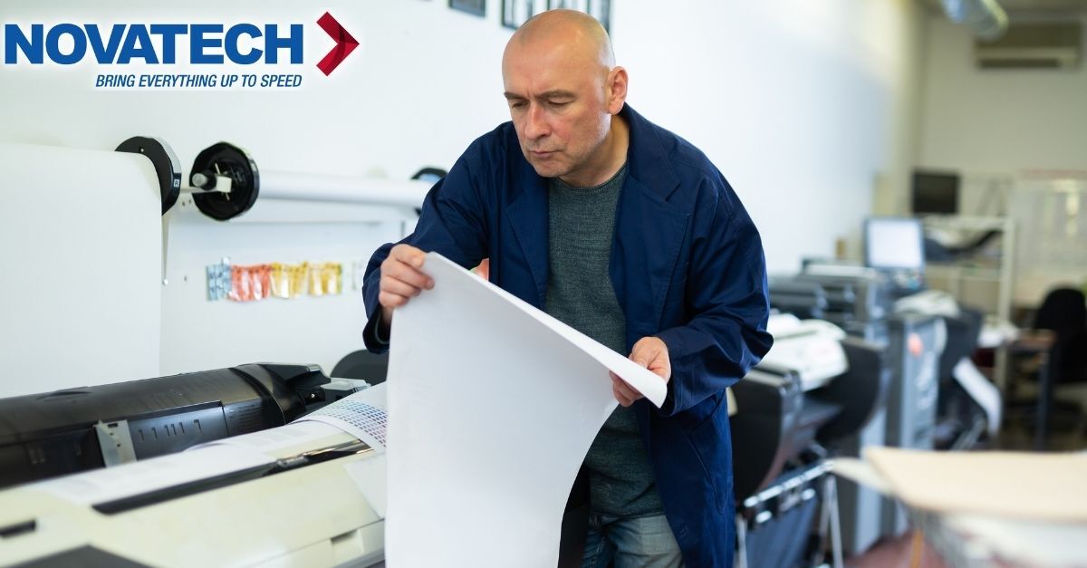A man in a blue lab coat inspects a large sheet of paper in a printing facility.