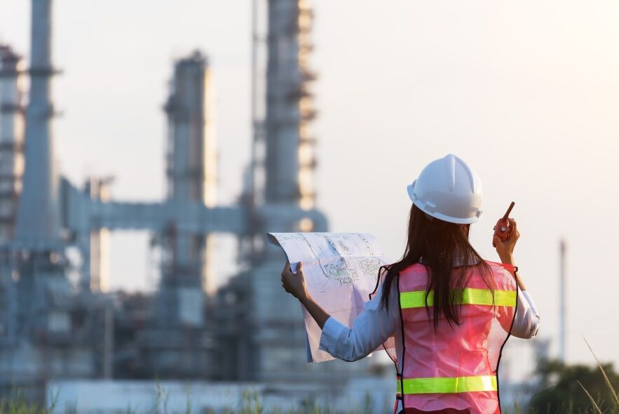 An engineer in a hard hat and safety vest reviews blueprints while holding a walkie-talkie in front of an industrial