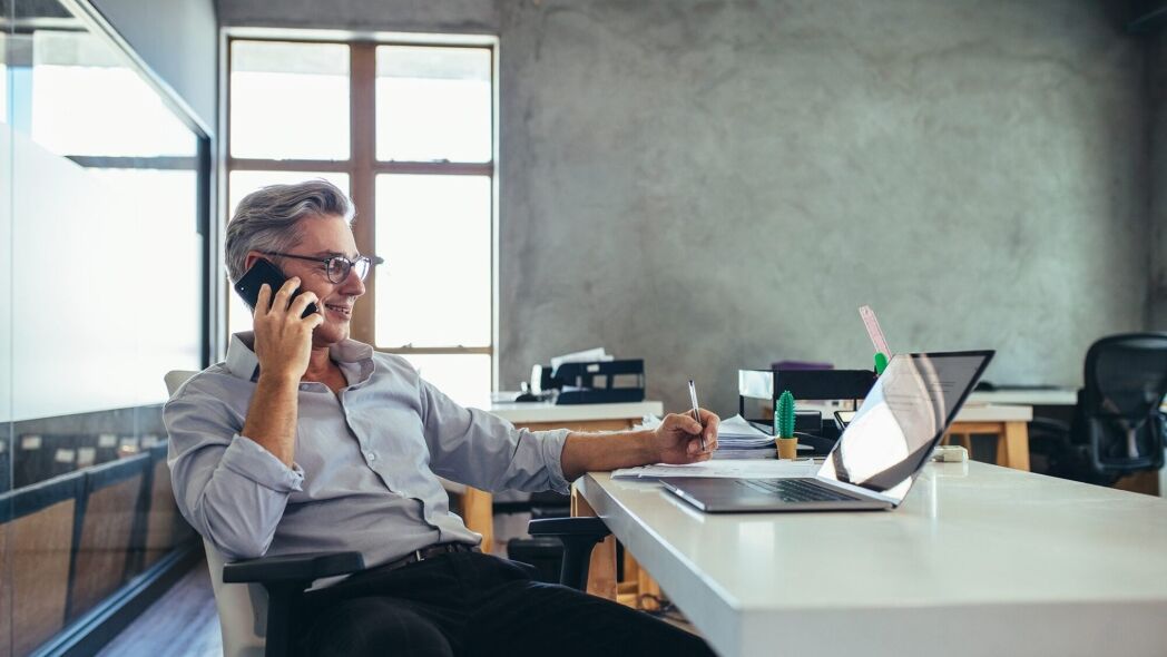 A businessman in glasses is on the phone and writing at his desk in a modern office.