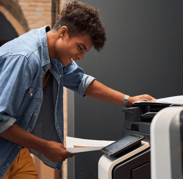 A young man in a denim jacket smiles as he places documents into a printer.