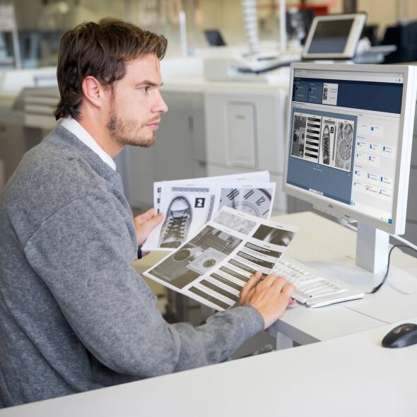 A man in a grey sweater reviews printed documents while working at a computer in an office setting.
