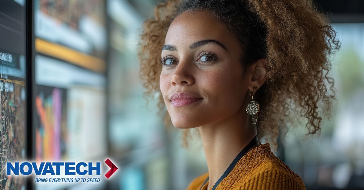 A woman with curly hair and freckles looks thoughtfully to the side in front of screens displaying data, with a company