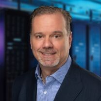 A smiling man with short brown hair and a goatee wears a blue pinstripe shirt and a dark suit jacket, with a blurred