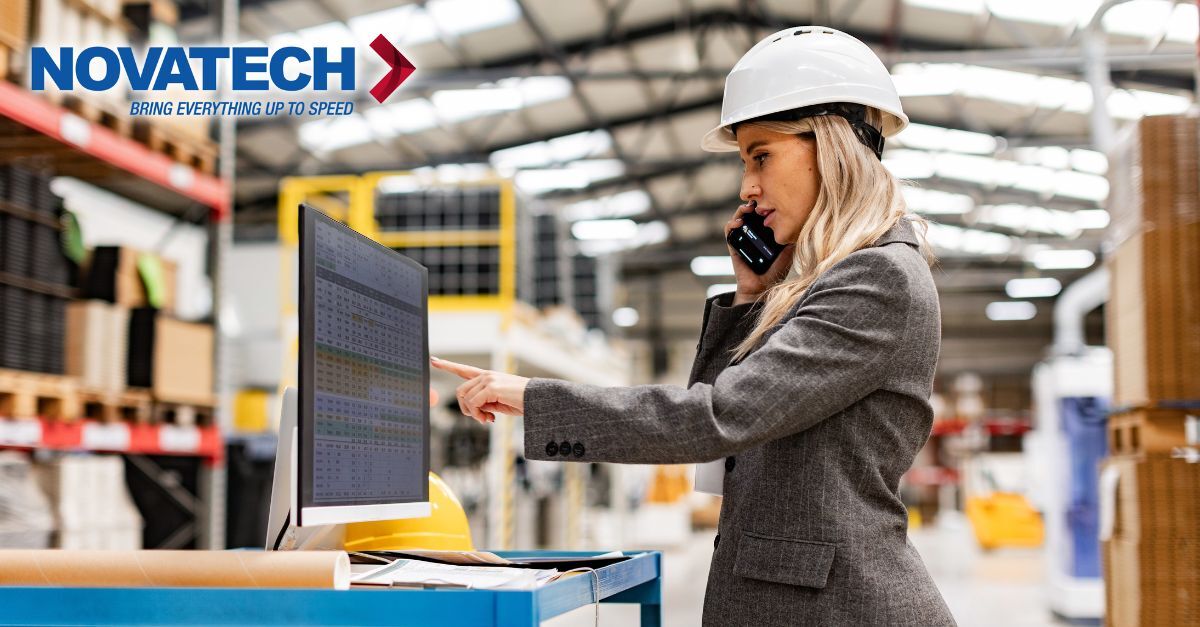 A woman in a hard hat and business attire talks on her phone while pointing at a computer screen in a warehouse.