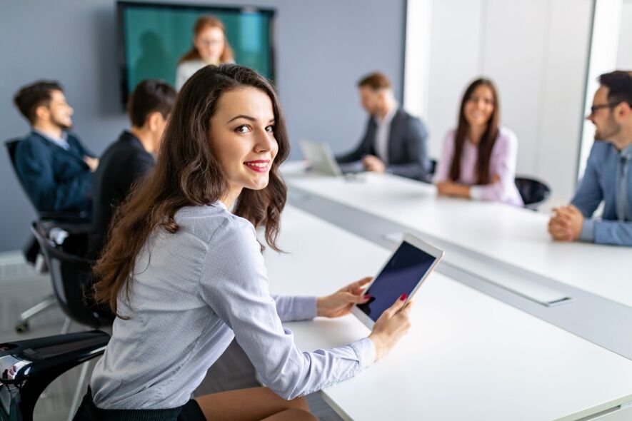 A young woman smiles at the camera while holding a tablet in a business meeting with colleagues in the background.