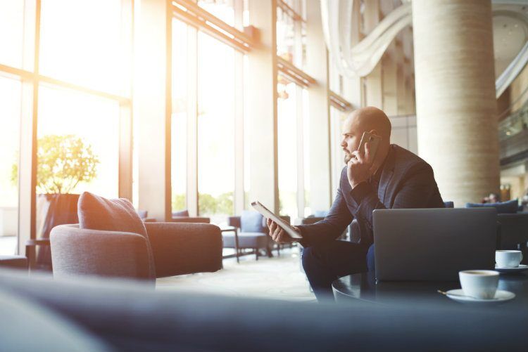 A businessman talks on his phone while holding a tablet in a modern lobby with large windows.