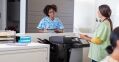 Two nurses interact at a reception desk with a tablet and printer.