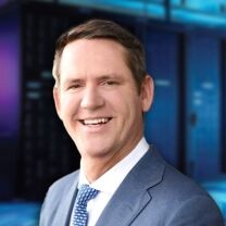 A smiling man in a blue suit and patterned tie is featured against a blurred background of a data center with blue and