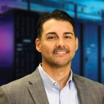 A man in a gray suit jacket and light blue collared shirt smiles at the camera with a blurred background of server