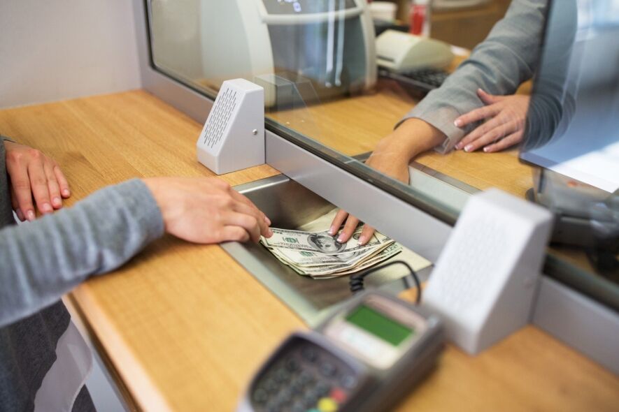 A bank teller hands a stack of US dollar bills to a customer through a security window.