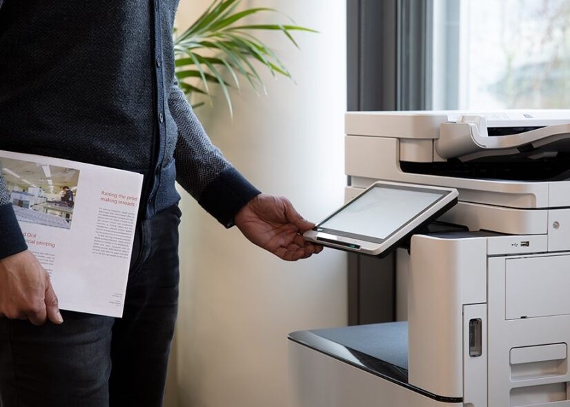 A person holding a brochure approaches a multifunction printer with a touchscreen interface.